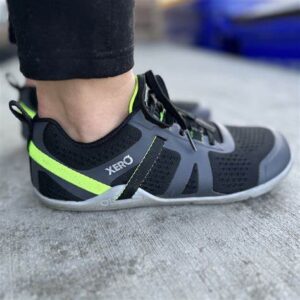 Close-up of a person wearing black and gray Xero barefoot shoes with yellow accents, showcasing comfort and style. The shoes are on a gray concrete surface, perfectly paired with black pants.