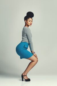 A person poses against a neutral background, wearing a striped top and blue skirt with high heels that accentuate their shoes. They have their hair in a high bun and are slightly bent forward, offering tips on striking the perfect pose while looking directly at the camera.