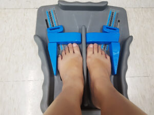 A pair of feet are positioned on a gray foot measurement device with blue sliding markers, resting on a light-colored floor. This tool ensures accuracy in determining shoe size, offering expert tips for the perfect fit every time.