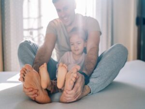 A man and a child sit on a bed, facing each other with their legs extended and feet touching—perhaps playfully comparing foot size or checking for signs of wide feet. The man is smiling, and both are wearing casual clothes. Sunlight filters through the window in the background.