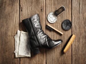 A black leather boot sits on a wooden surface, accompanied by an array of shoe polishing tools. Among them are two types of shoe brushes essential for maintaining shine, alongside a soft cloth and tins of white and black polish.