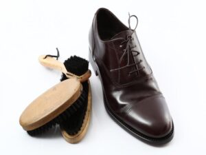 A polished brown leather dress shoe with laces is placed next to two wooden-handled shoe brushes with black bristles, illustrating the importance of understanding shoe brush types. The shoe rests elegantly on a white surface.