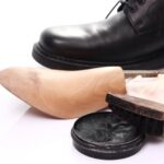 A black leather shoe is placed next to a spiral shoe tree, a shoe brush, a cloth, and an open tin of black shoe polish on a white surface.