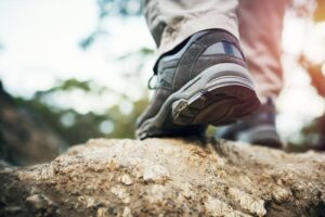 Close-up of a person wearing hiking shoes, walking on a rocky path. The focus is on the new shoes and terrain, with sunlight filtering through trees in the background. Perfect conditions shouldn't distract from the durability required for such adventurous strolls.