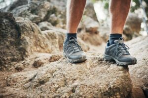 A person stands on a rocky trail in new gray hiking shoes and black socks. The scene focuses on their legs and feet, capturing the texture of the rocks and wondering when to condition them for optimal performance in this rugged outdoors environment.