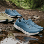 Three pairs of Xero Shoes in green, light blue, and dark blue rest on rocky ground near a shallow stream, with a forested backdrop—showcasing their durability and performance in the great outdoors.