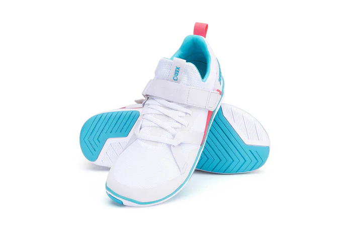 A pair of white athletic sneakers, the Forza Trainer by Xero Shoes, features light blue soles, a blue logo, and pink accents. One shoe stands upright while the other lies on its side, showcasing the tread pattern of this performance footwear.