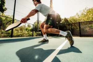 A person in athletic clothing and pickleball shoes is playing pickleball outdoors on a sunny day. They are holding a paddle and are in a low, athletic stance on a blue court with a white dividing line, with trees and a fence in the background.