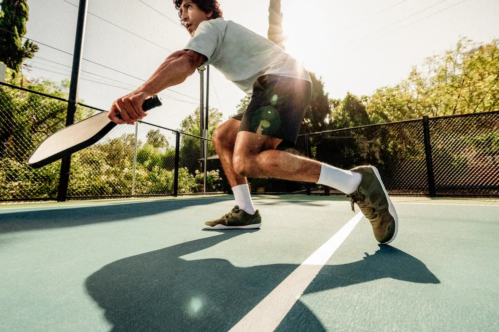A person in athletic clothing and pickleball shoes is playing pickleball outdoors on a sunny day. They are holding a paddle and are in a low, athletic stance on a blue court with a white dividing line, with trees and a fence in the background.