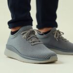 A person wearing dark pants and light gray Nexus Knit minimalist footwear by Xero Shoes with white soles, standing on a light surface. The shoes have laces and a textured pattern.