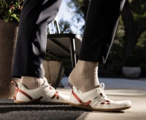 A person wearing beige socks and white Xero Shoes Prio Coast with red accents is stepping on the heel of one slip-on shoe outdoors, next to a chair and a planter, as sunlight casts shadows on the ground.