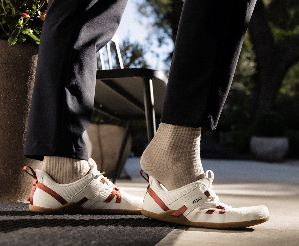 A person wearing beige socks and white Xero Shoes Prio Coast with red accents is stepping on the heel of one slip-on shoe outdoors, next to a chair and a planter, as sunlight casts shadows on the ground.