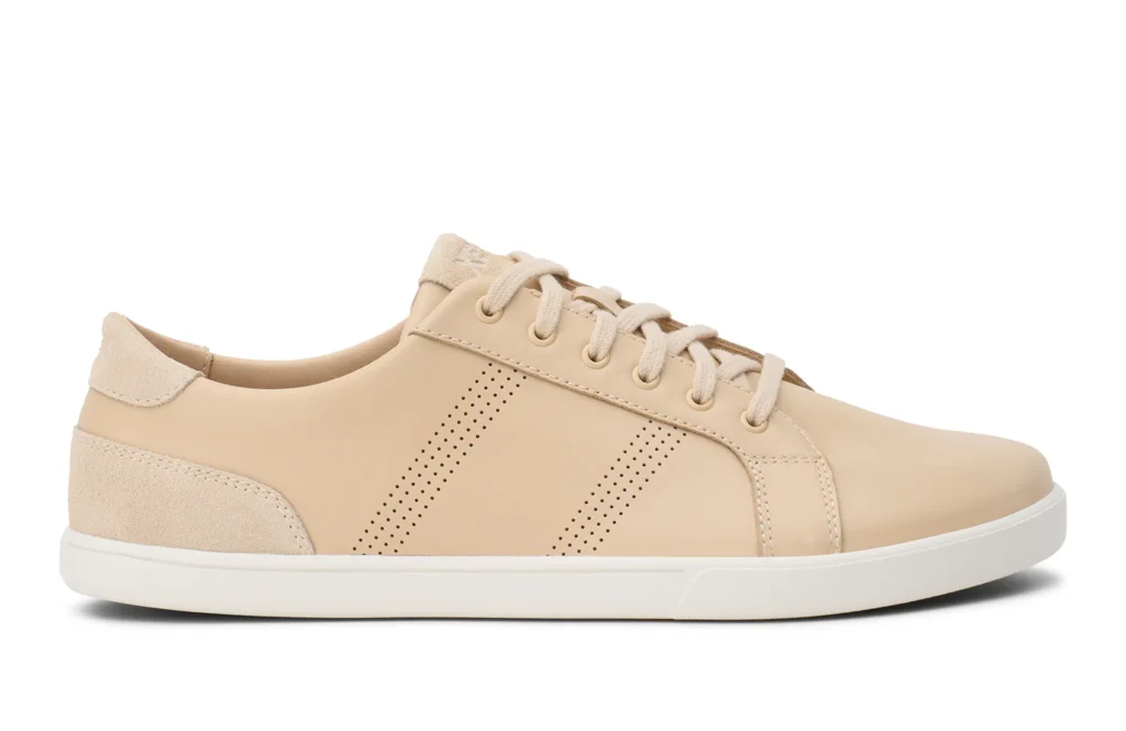 A beige low-top sneaker from Xero Shoes with a white sole, beige laces, suede detailing at the heel, and perforated side stripes. The shoe is photographed from the side on a white background.