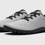 A pair of light gray, lace-up athletic shoes from Xero Shoes, featuring black soles, black laces, black inner lining, and a small circular logo on the heel area—showcasing quality craftsmanship on a plain light background.