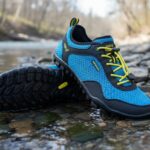 A pair of blue and black Xero Shoes Aqua X Sport with yellow laces are placed on rocks beside a river, one standing and the other leaning. The forested riverside backdrop highlights these adventure footwear essentials.