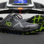 A pair of gray and black athletic running shoes with green and blue accents are placed on the belt of a treadmill, making them perfect for indoor workouts or treadmill running, with the digital display and control panel visible in the background.