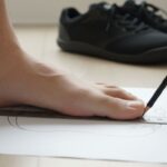 A person traces the outline of their foot on a sheet of paper using a pencil and ruler to measure for Xero Shoes, with black shoes in the background on a light wooden floor.