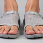 Close-up of two feet wearing gray knit open-toe Xero Shoes sandals with adjustable straps and the word "Xero" on each strap, standing on a light gray surface. Designed with wide toe boxes, these sandals provide extra comfort—perfect for those with bunions. Toes are visible through the open front.