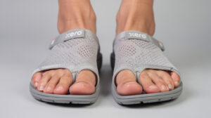 Close-up of two feet wearing gray knit open-toe Xero Shoes sandals with adjustable straps and the word "Xero" on each strap, standing on a light gray surface. Designed with wide toe boxes, these sandals provide extra comfort—perfect for those with bunions. Toes are visible through the open front.